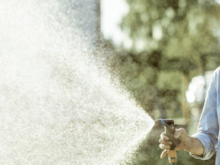Man around 30 years old smiling while watering his garden with a hose. The water is spraying towards the camera.