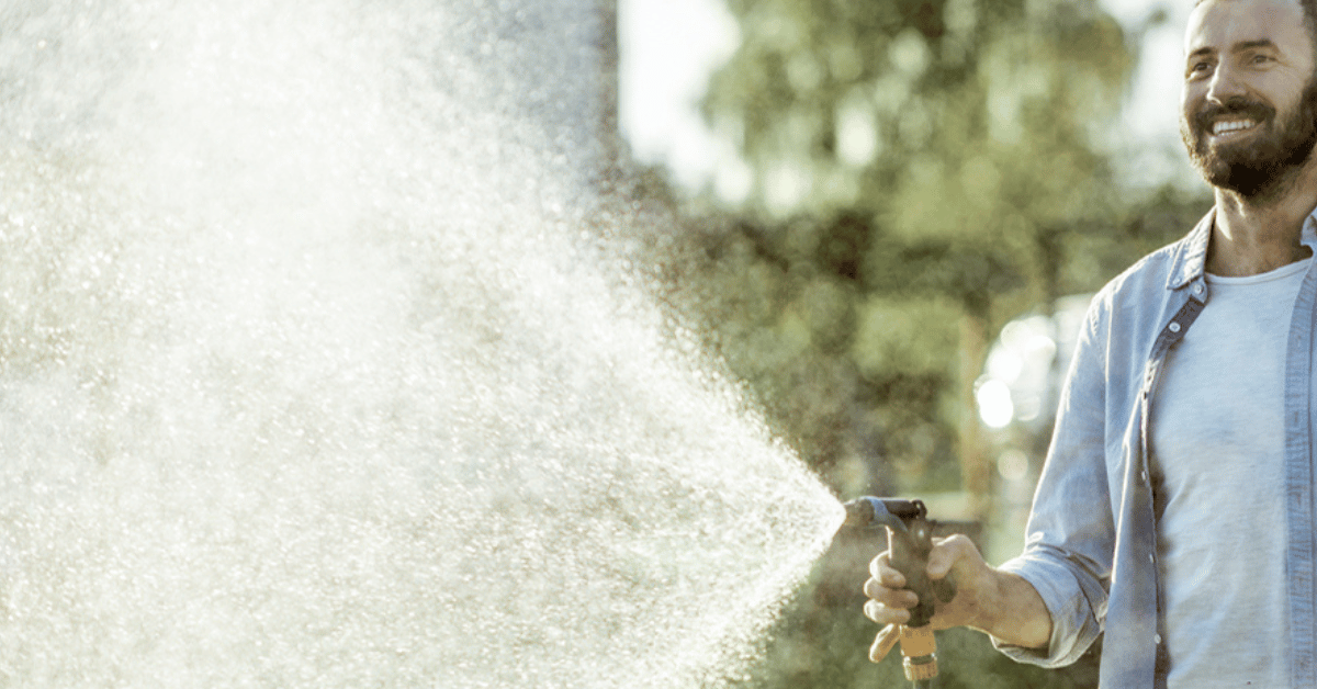 Man around 30 years old smiling while watering his garden with a hose. The water is spraying towards the camera.